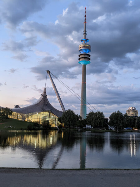Olympiapark München: Blick auf den Olympiaturm und den Olympiasee in der Abenddämmerung. Im Hintergrund ist das Schwimmbad und das BMW Gebäude zu sehen.
